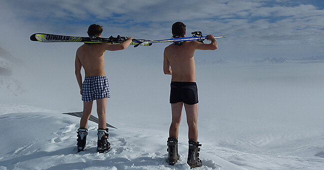 zwei junge Männer in Unterhosen und Skier tragend auf Schneegipfel