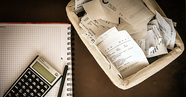 Papierkorb gefüllt mit Papierblättern, links ein Block mit Stift und Taschenrechner